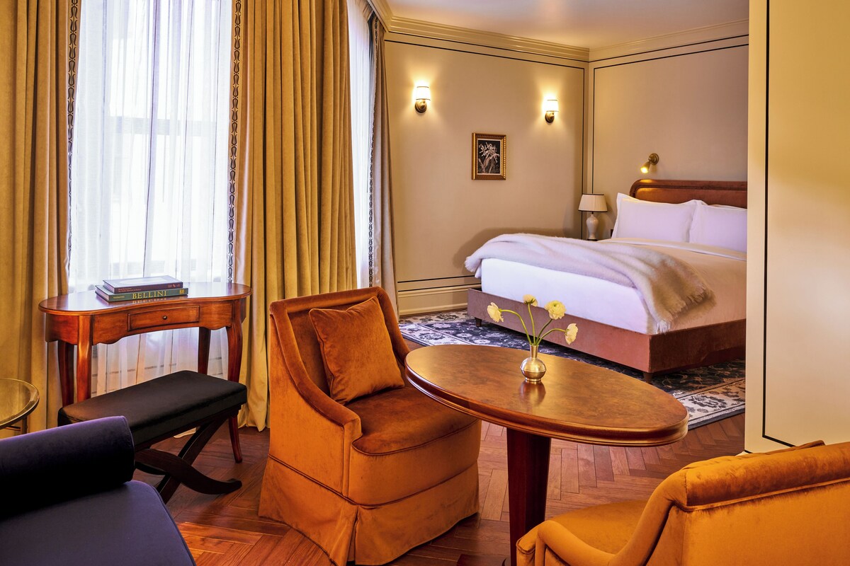 A hotel room with a bed, a desk, two armchairs, a small table, and a vase with flowers.