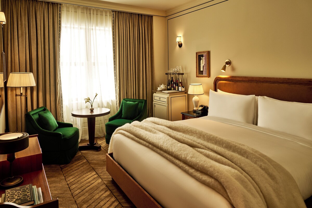A hotel room with a large bed, two chairs, a small table, and a desk with a lamp.