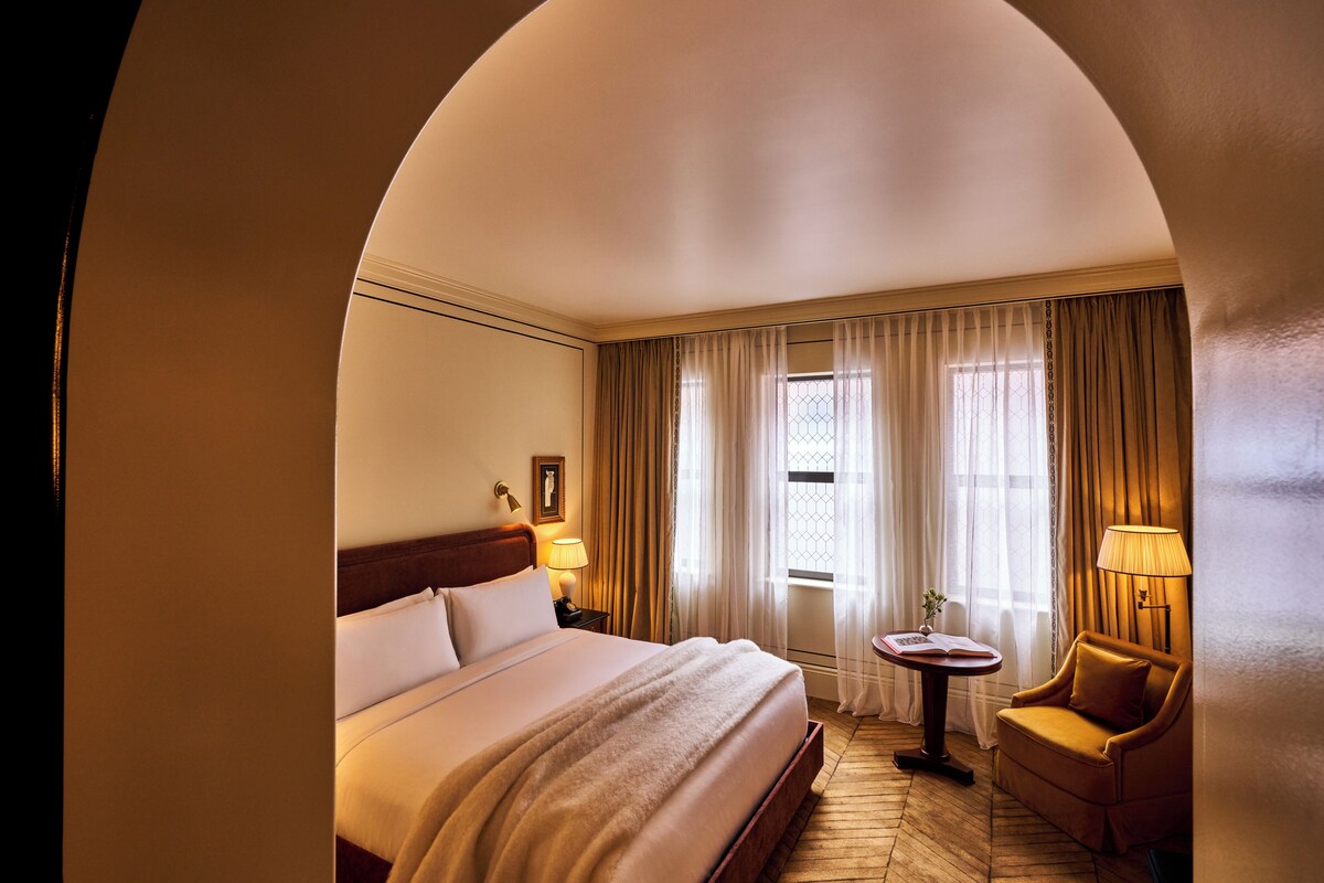 A hotel room with a large bed, a small table with a lamp, a chair, and a window with curtains.
