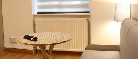 A room with a wooden floor, a round table with a remote control, a sofa, a window with blinds, a radiator, and a lamp.