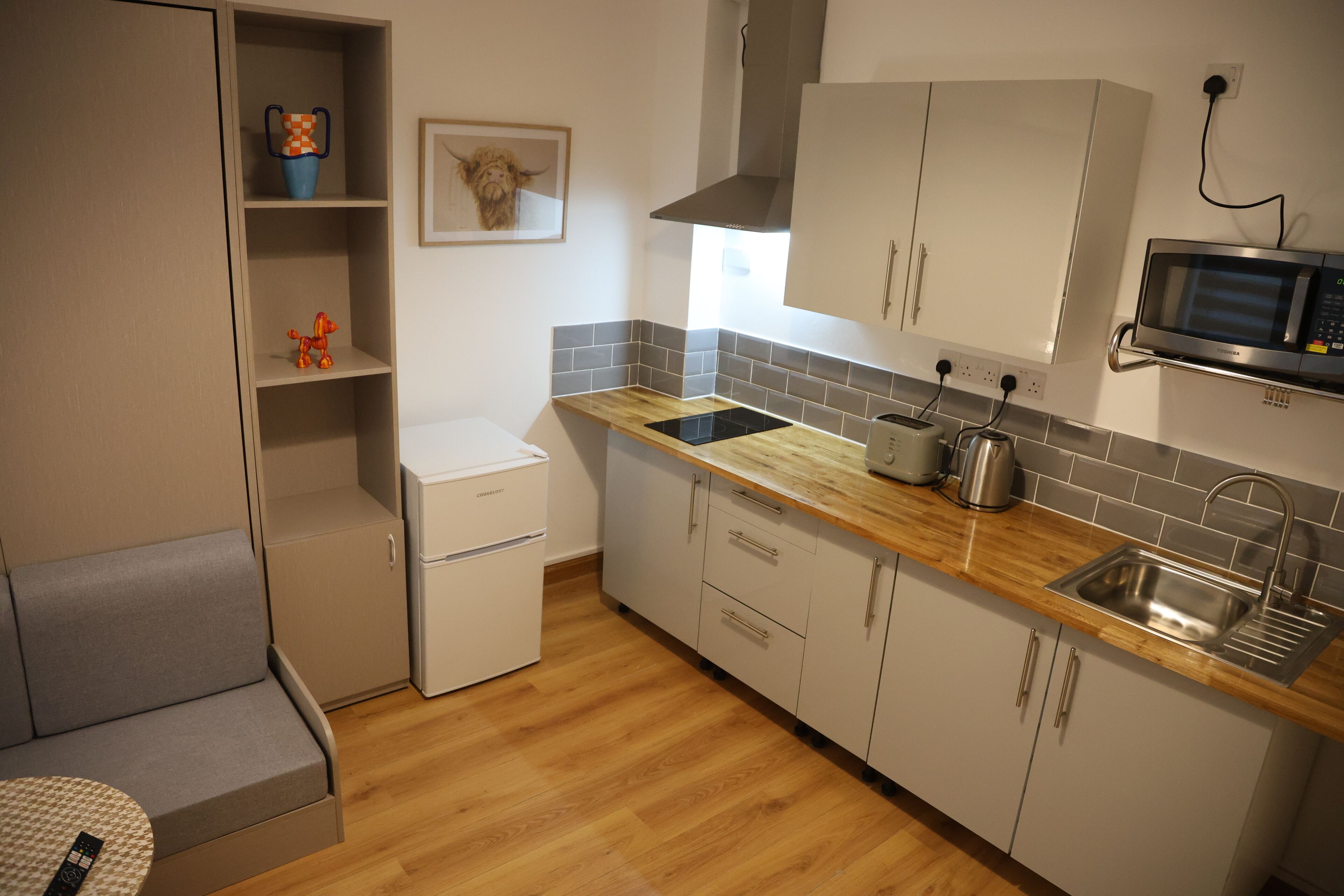 Een kleine keuken met een ingebouwde magnetron, een broodrooster en een spoelbak. Ook is er een kleine koelkast en een houten aanrecht.