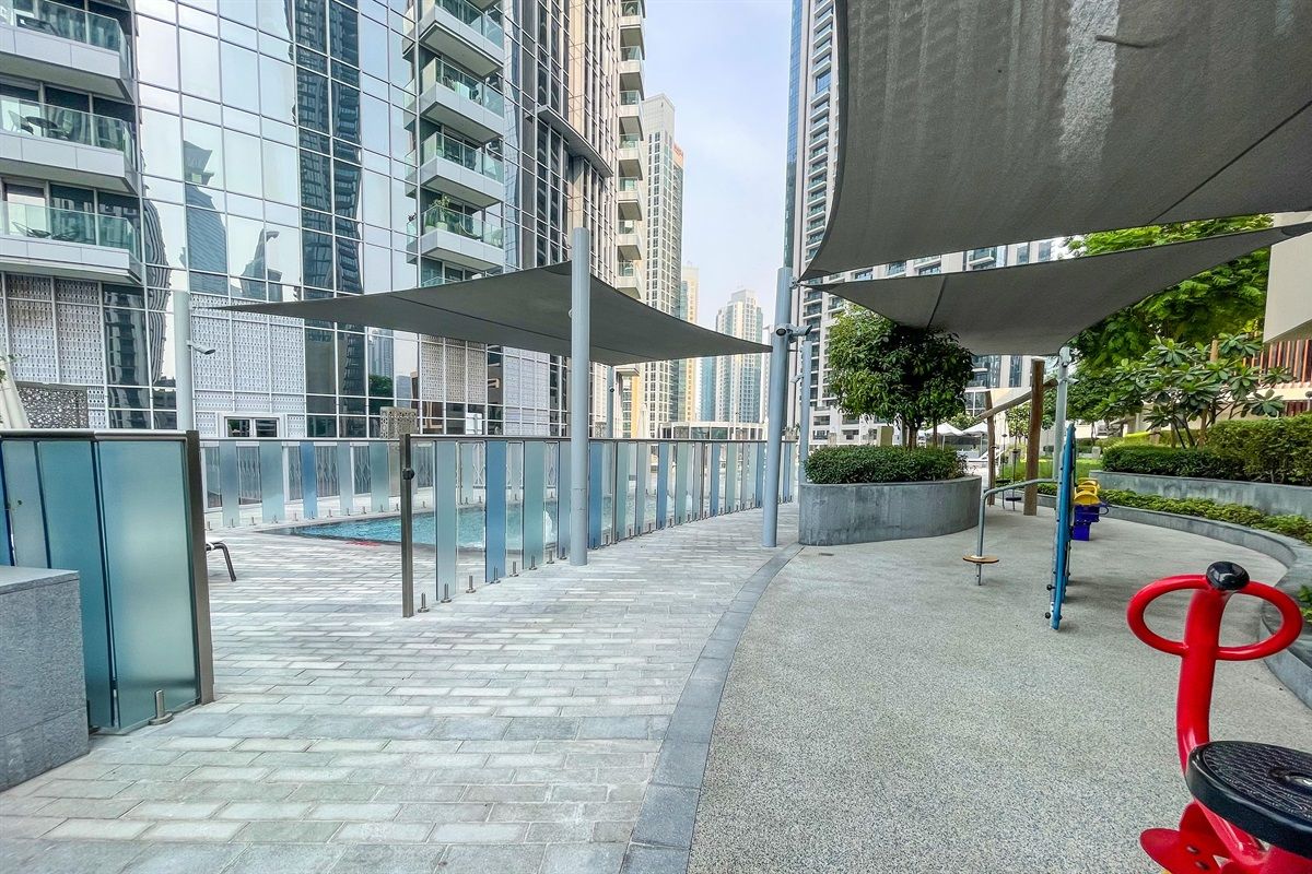 A modern outdoor area with a paved walkway, a children's play area, and high-rise buildings in the background.
