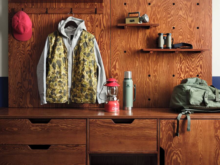 Une commode en bois, un tapis à carreaux, une paire de bottes, une bouilloire rouge, un thermos vert, une veste de camouflage et une casquette rouge.