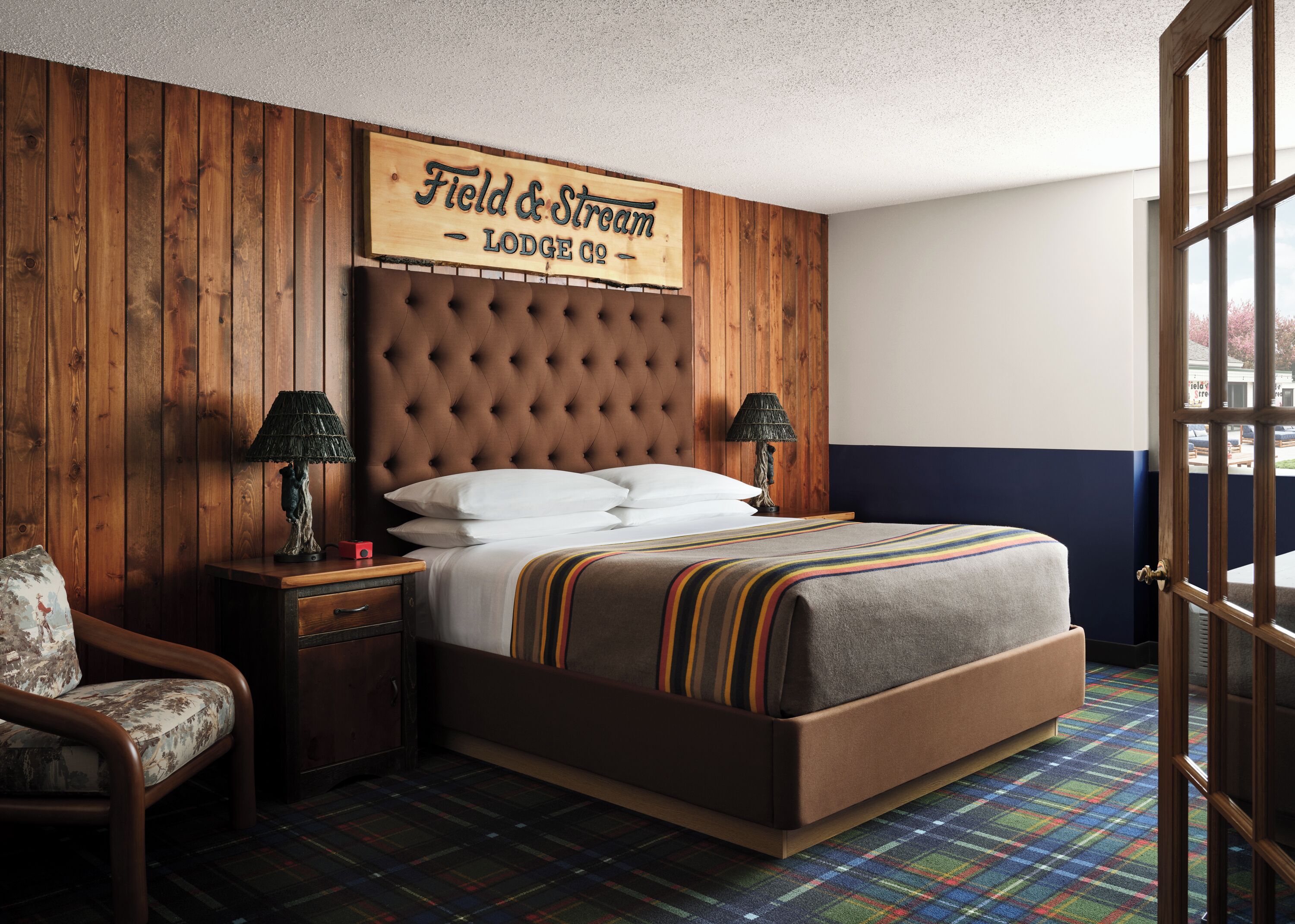 A bedroom with a tufted headboard, a bed with white linens, striped bedding, a nightstand with lamps, and a plaid-patterned carpet.