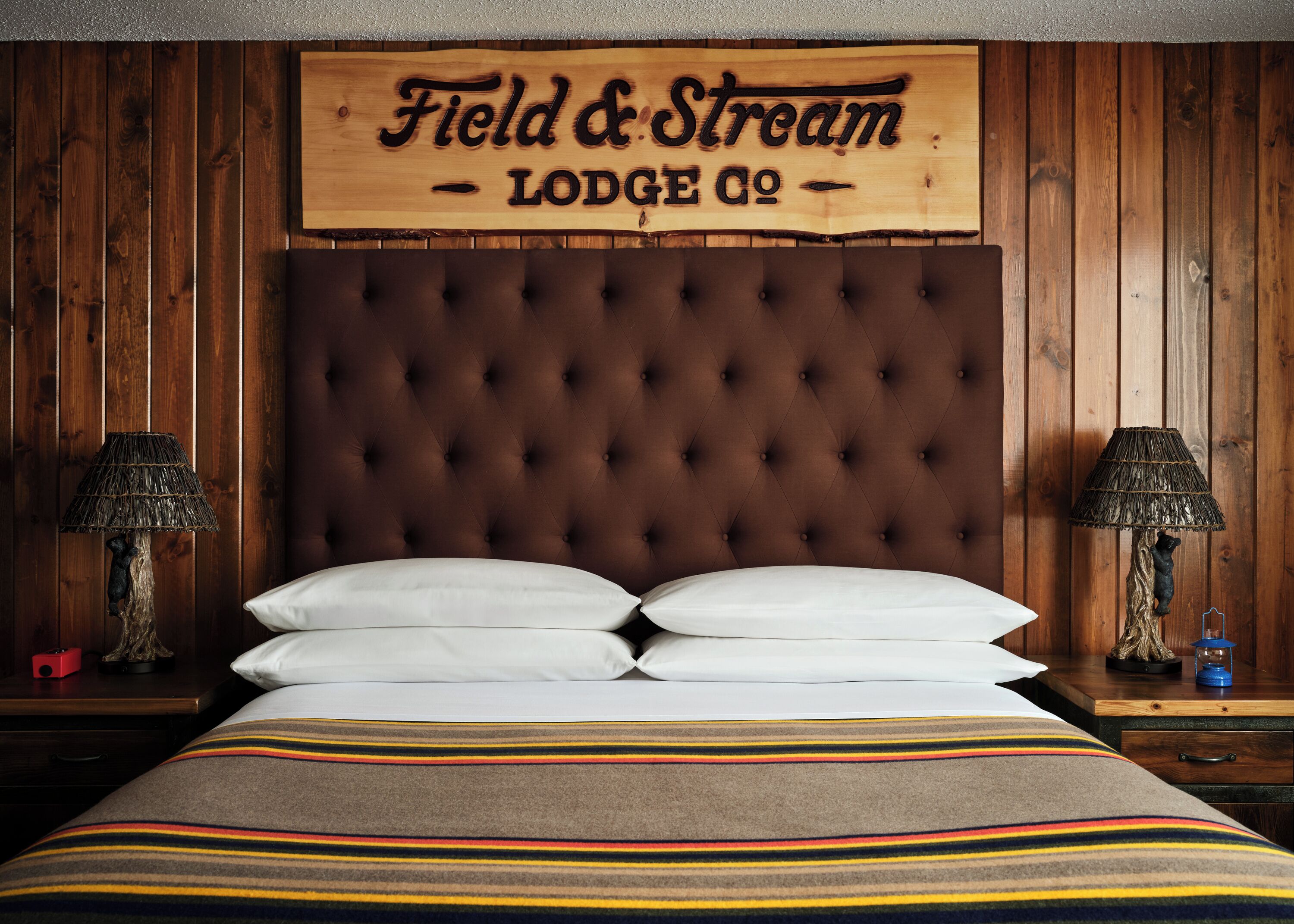 Ein Schlafzimmer mit einer gepolsterten Kopfteile, zwei Nachttischlampen, einer gestreiften Decke und einem Holzschild über dem Bett mit der Aufschrift 'Field & Stream Lodge Co.'