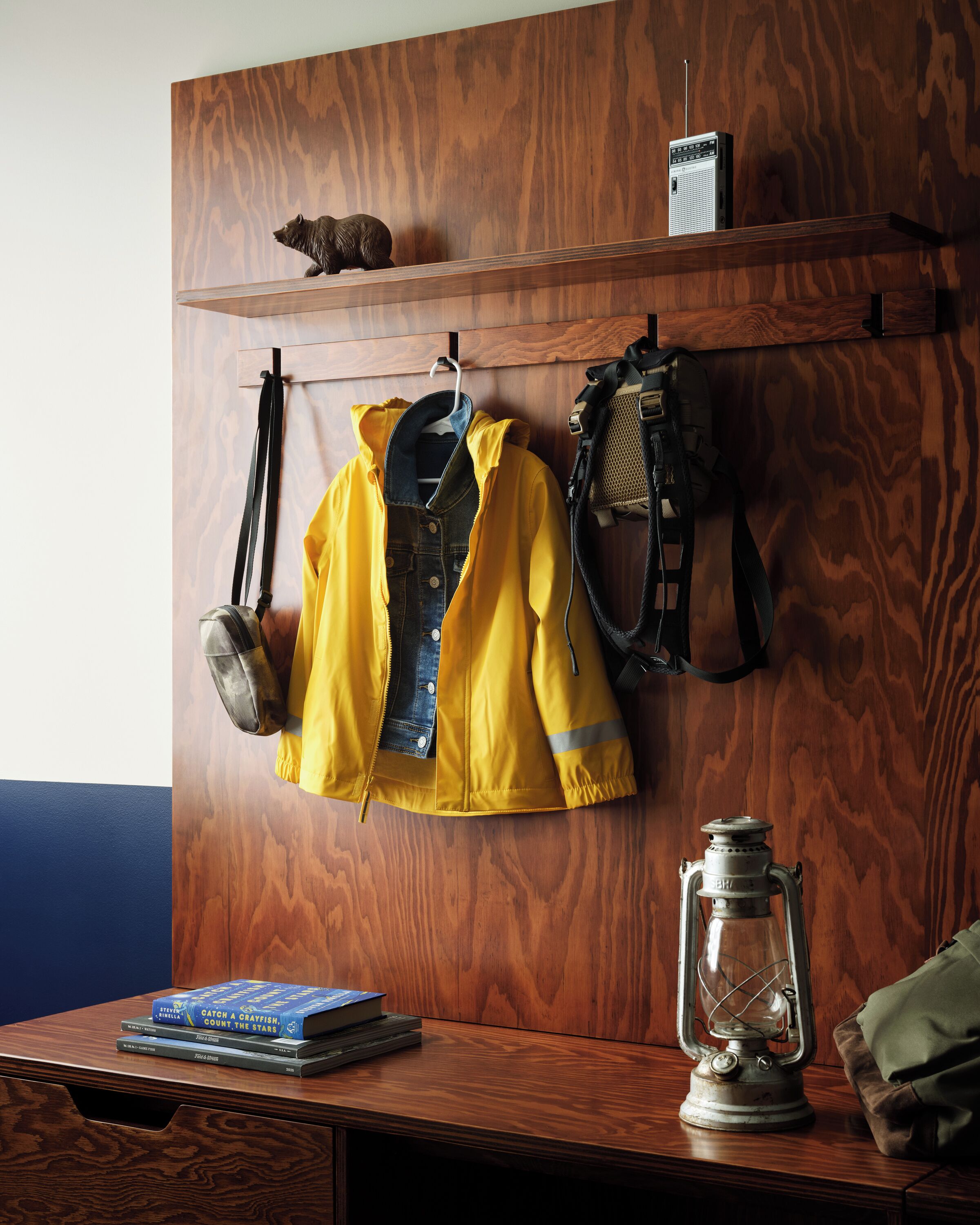 Una pared de madera con un estante que sostiene una radio, una pequeña figurilla y un farol. Abajo, una chaqueta amarilla, un chaleco vaquero y una mochila están colgados de ganchos. Un libro y un farol se encuentran sobre una mesa de madera.