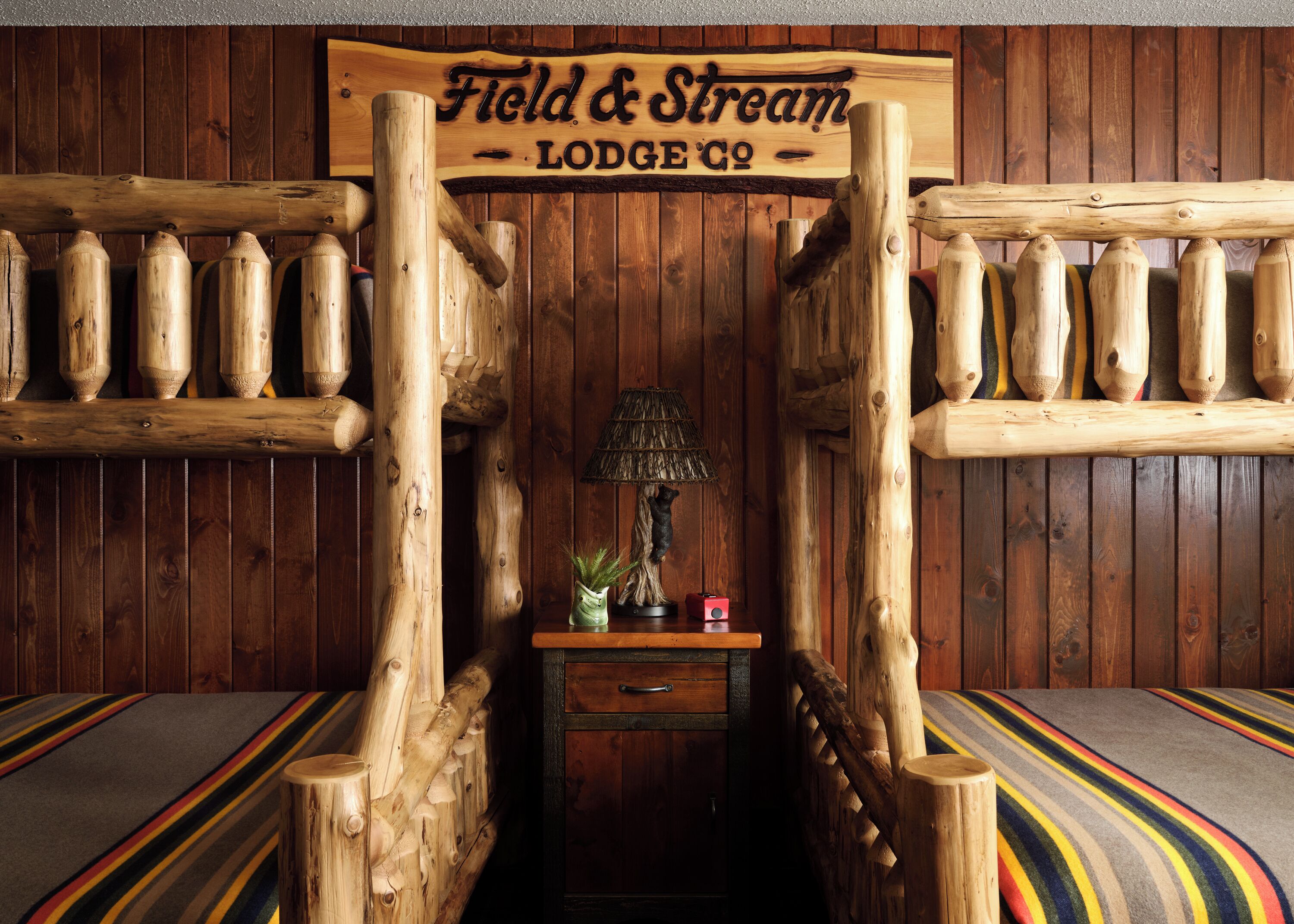 Een houten kamer in blokhutstijl met een bord waarop 'Field & Stream Lodge Co.' staat, en een kleine tafel met een lamp en decoratieve objecten.