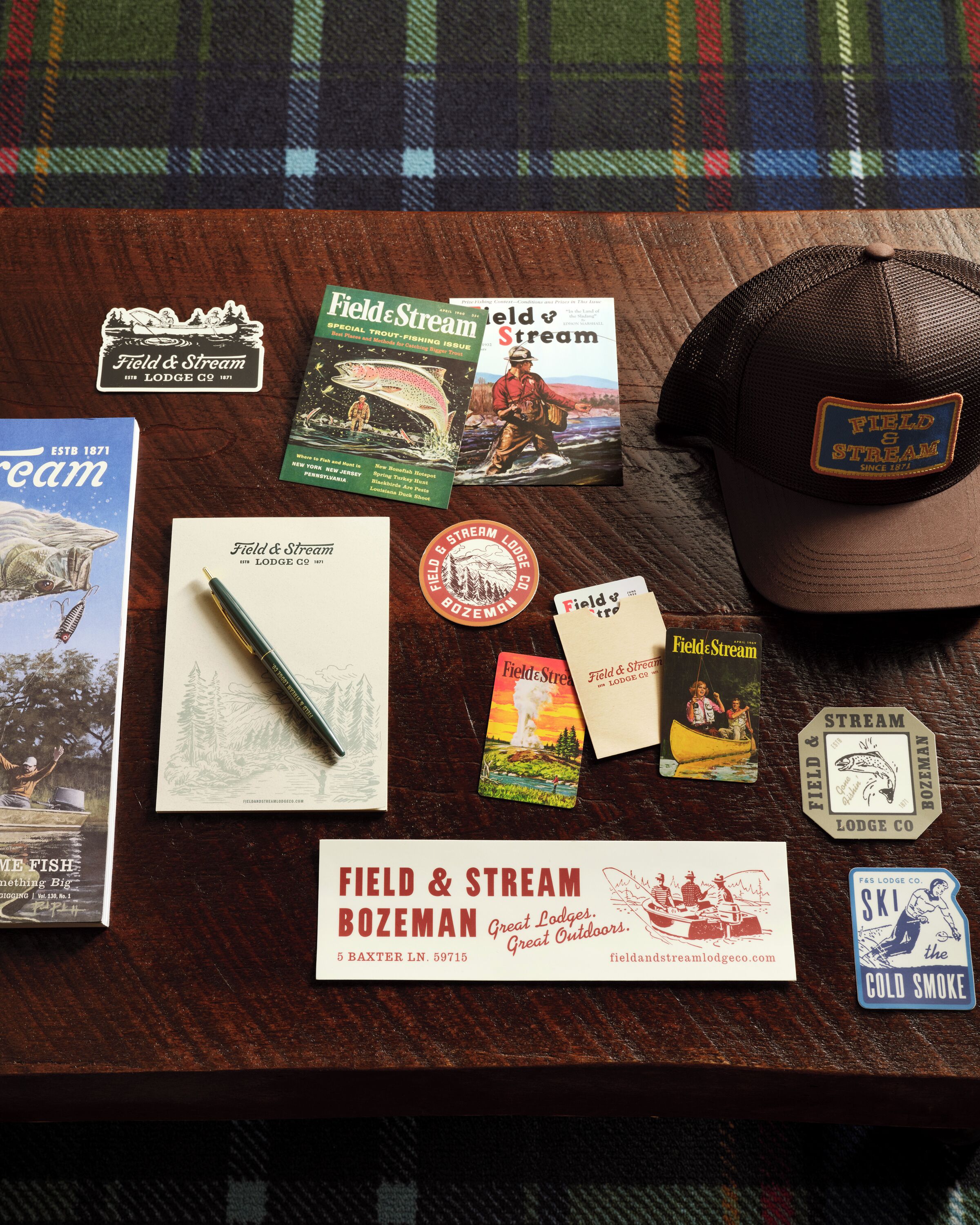 A wooden table with various items including a Field & Stream Lodge Co. cap, a pen, postcards, and a book titled 'Field & Stream'.