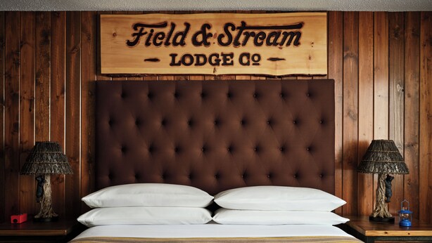 A room with a tufted headboard, two bedside lamps, and a bed with white pillows. A sign above reads 'Field & Stream Lodge Co.'