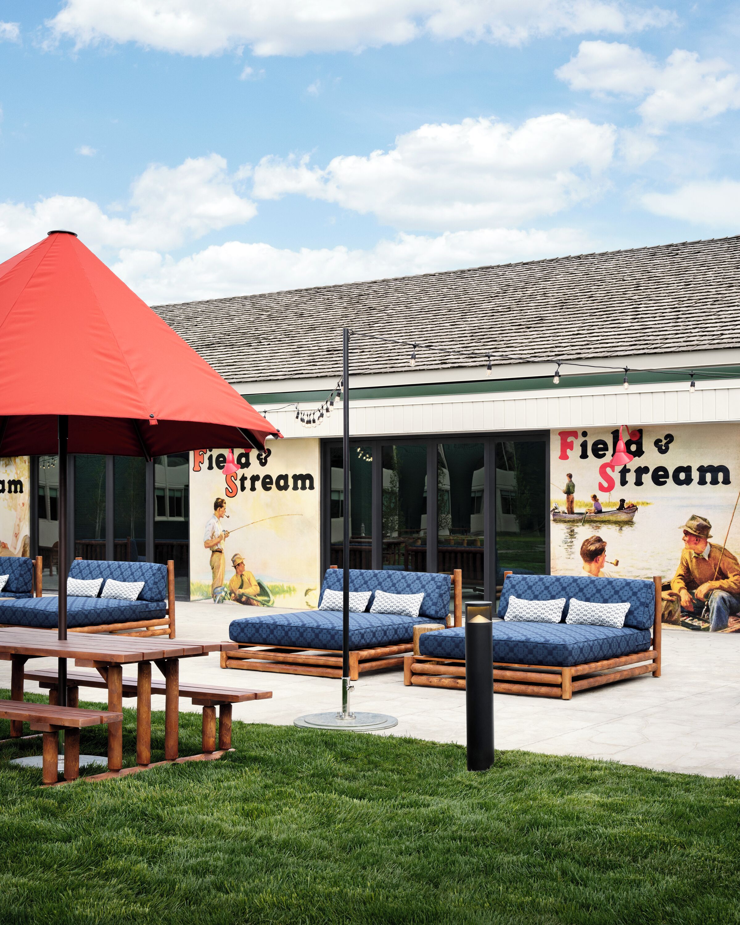 Een terras met lounge fauteuils en een rode parasol, een muurschildering met visscènes, en een gebouw met een pannendak.