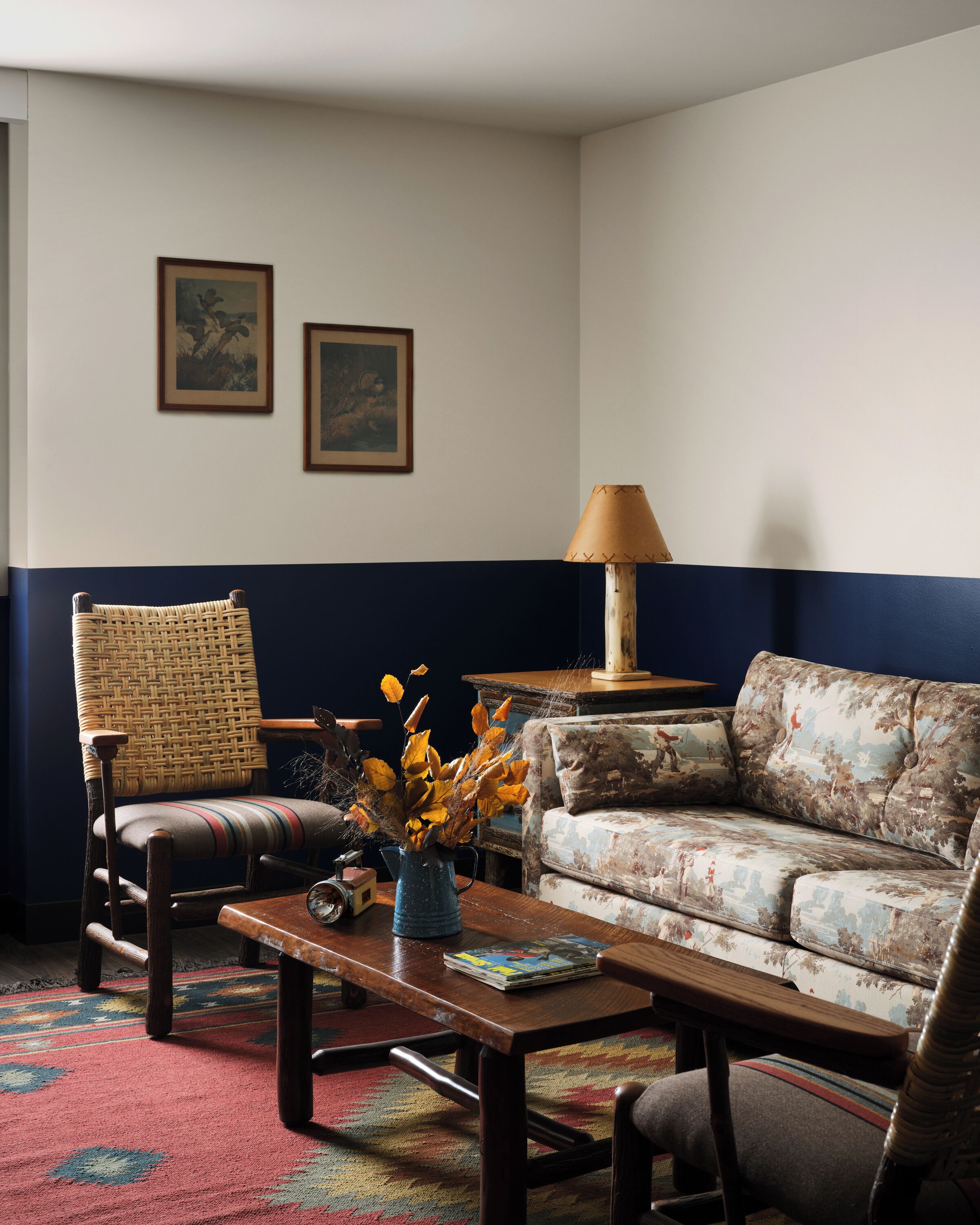 A living room with a patterned sofa, a wicker chair, a coffee table with a vase of dried flowers, and a lamp.