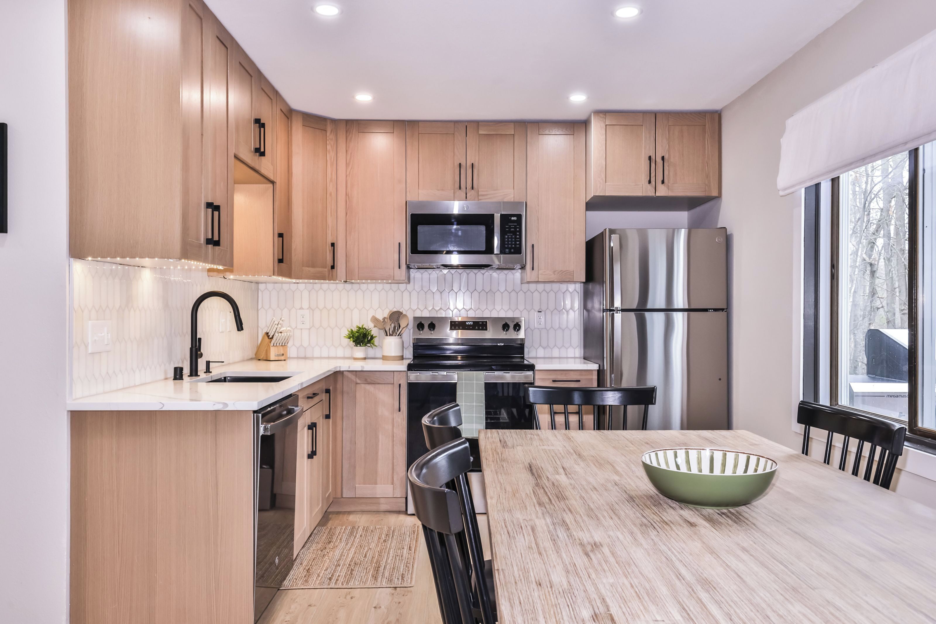 A modern kitchen with wooden cabinets, stainless steel appliances, a dining table with chairs, and a large window.