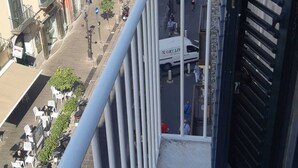 Un balcón con vista a una calle flanqueada por edificios, una furgoneta estacionada y mobiliario exterior.