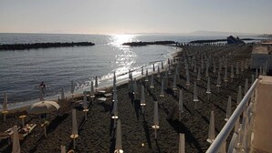 Una playa con hileras de sombrillas y sillas de descanso, un muelle al fondo, y el sol reflejándose en el agua.