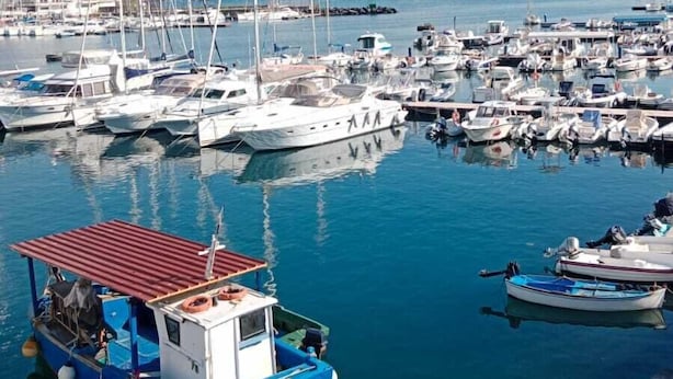Un puerto deportivo con varias embarcaciones amarradas, un bote azul con techo rojo en primer plano, y un paisaje montañoso al fondo.