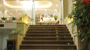 Zona de recepción de un hotel con una escalera que conduce al piso superior, una mesa de recepción con un reloj y plantas en macetas.