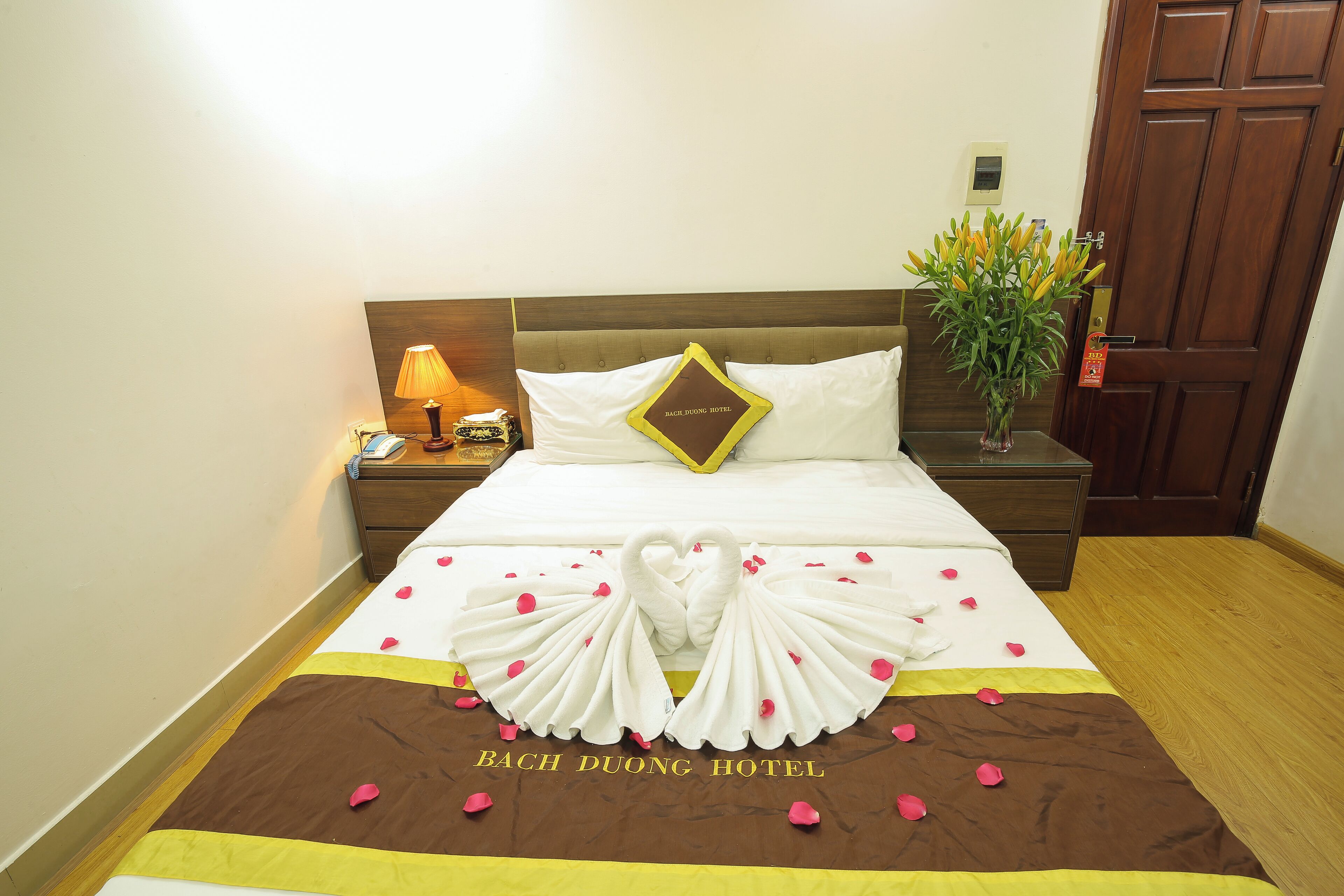 A hotel room with a neatly made bed, a bedside lamp, a nightstand, and a potted plant.