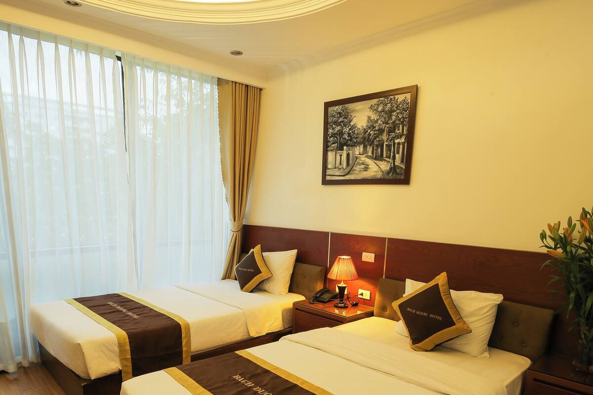 A hotel room with two beds, a nightstand with a lamp, a framed picture on the wall, and a large window with curtains.