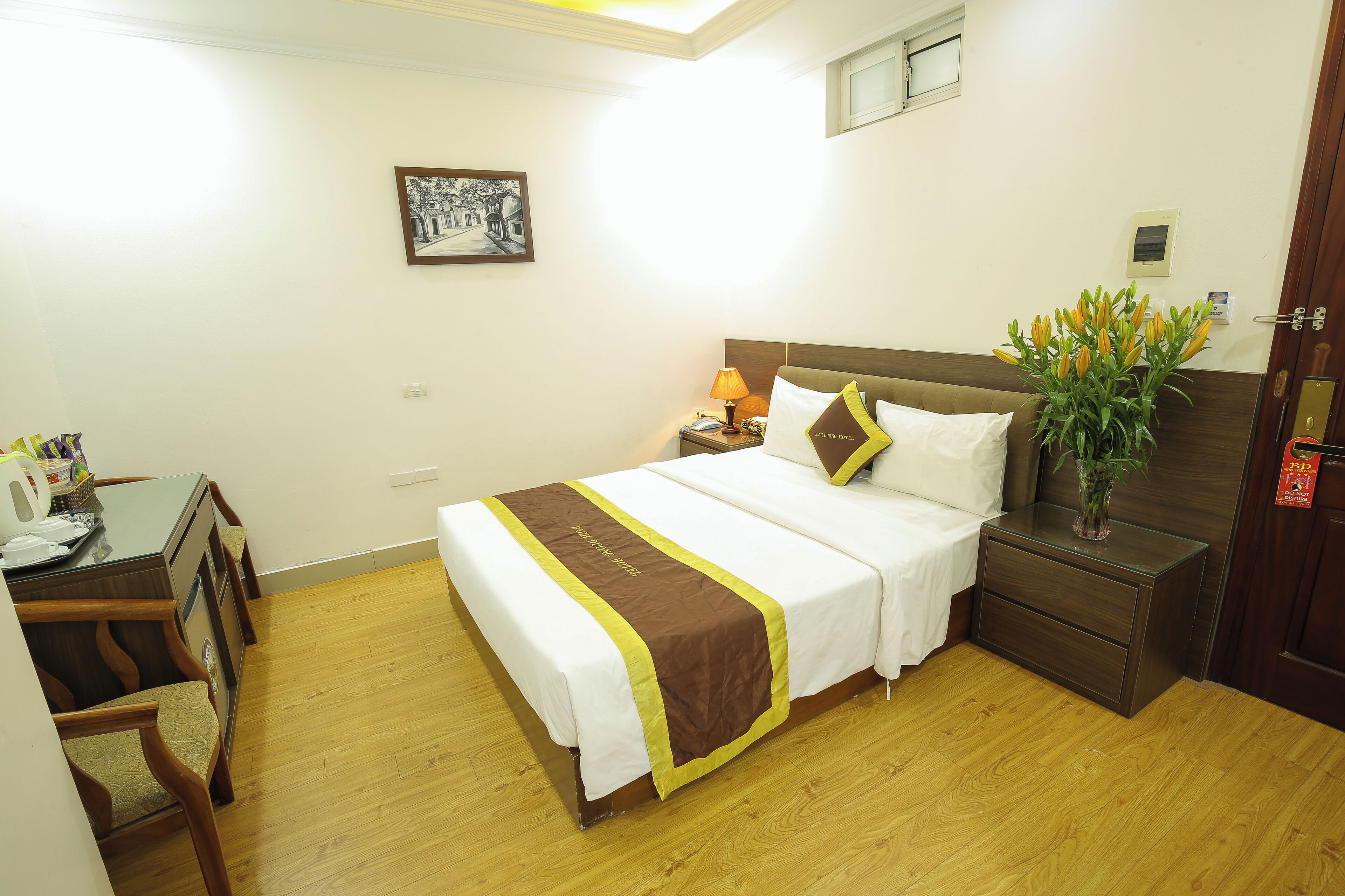 Una habitación de hotel con cama, mesita de noche, lámpara y un jarrón con flores.