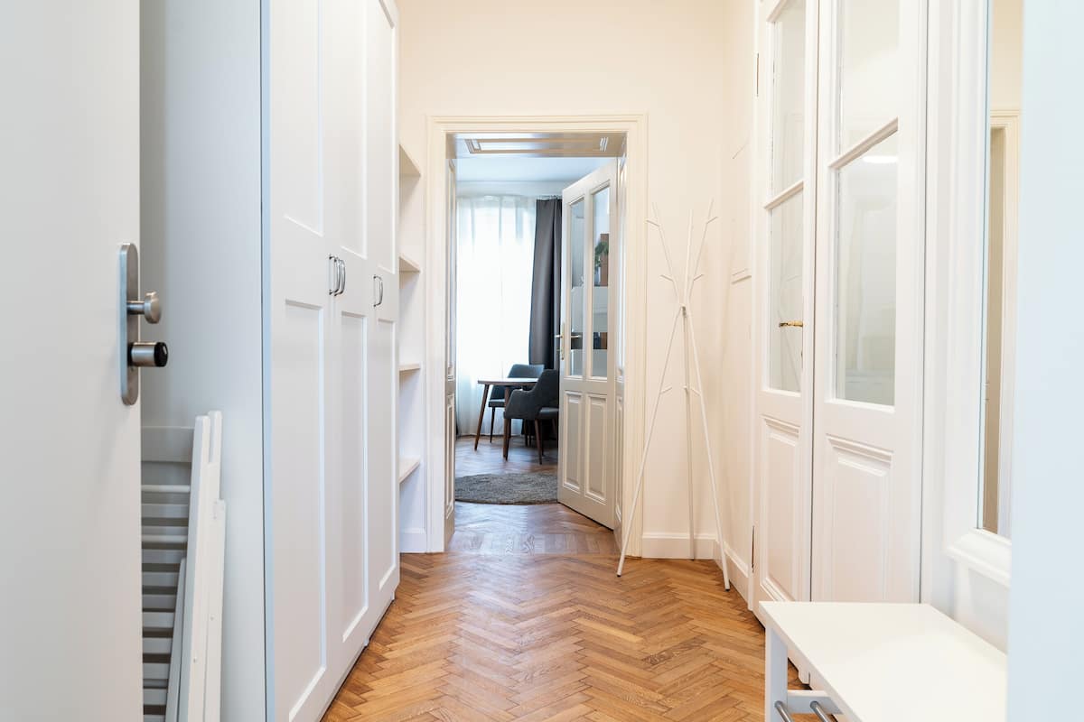 A hallway with white doors, a herringbone wooden floor, and a view into a dining area with a table and chairs.