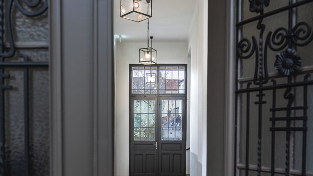 Un corridoio con una porta in legno scuro, una porta vetrata con un oblò e un lampadario dal design geometrico.