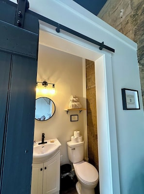 A bathroom with a white sink, a toilet, and a mirror. There is a shelf with towels and a framed picture on the wall. - The Stonehouse Makers' Maisonette (Pictou)