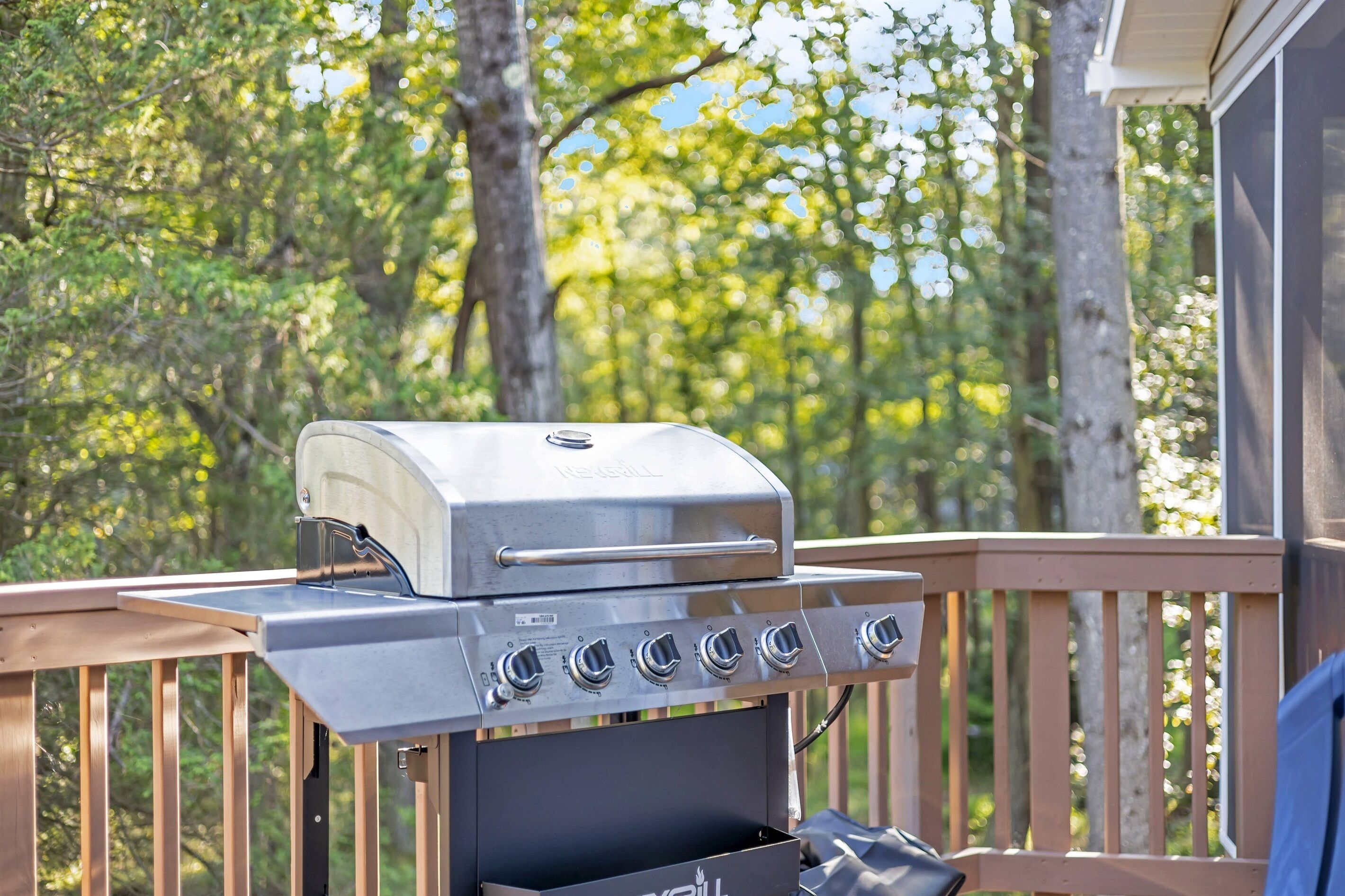 A stainless steel grill with four burners on a wooden deck, surrounded by trees.