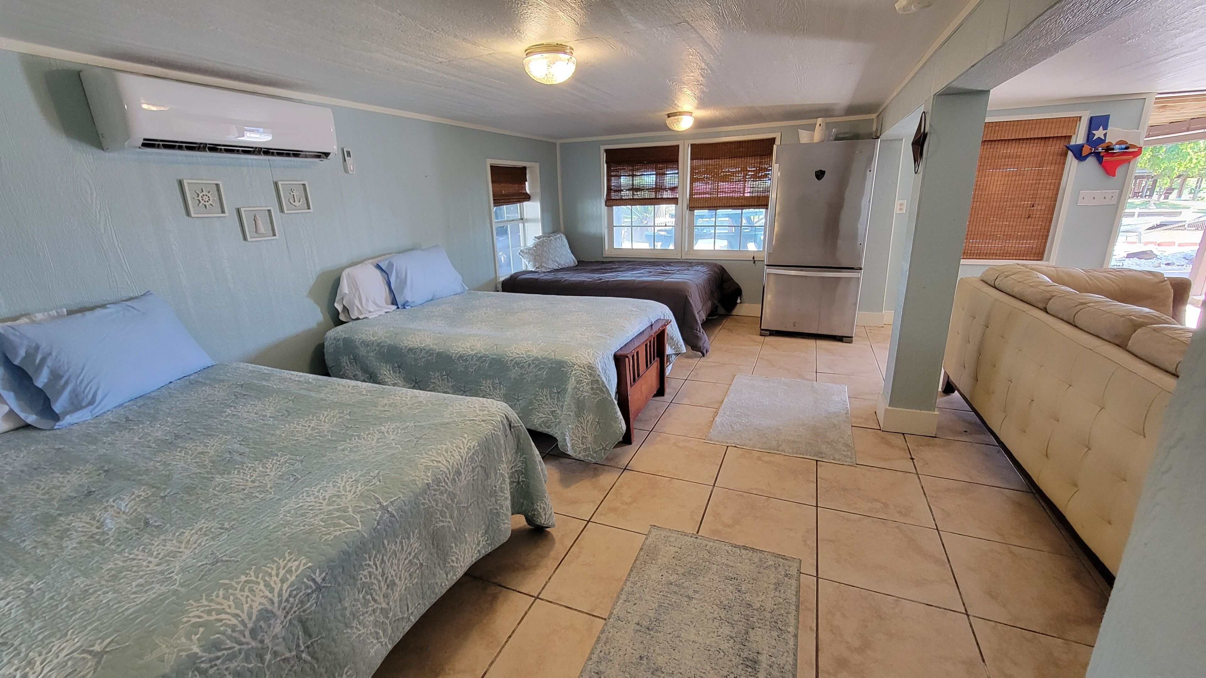 A room with two beds, a sofa, a refrigerator, and a window with blinds.