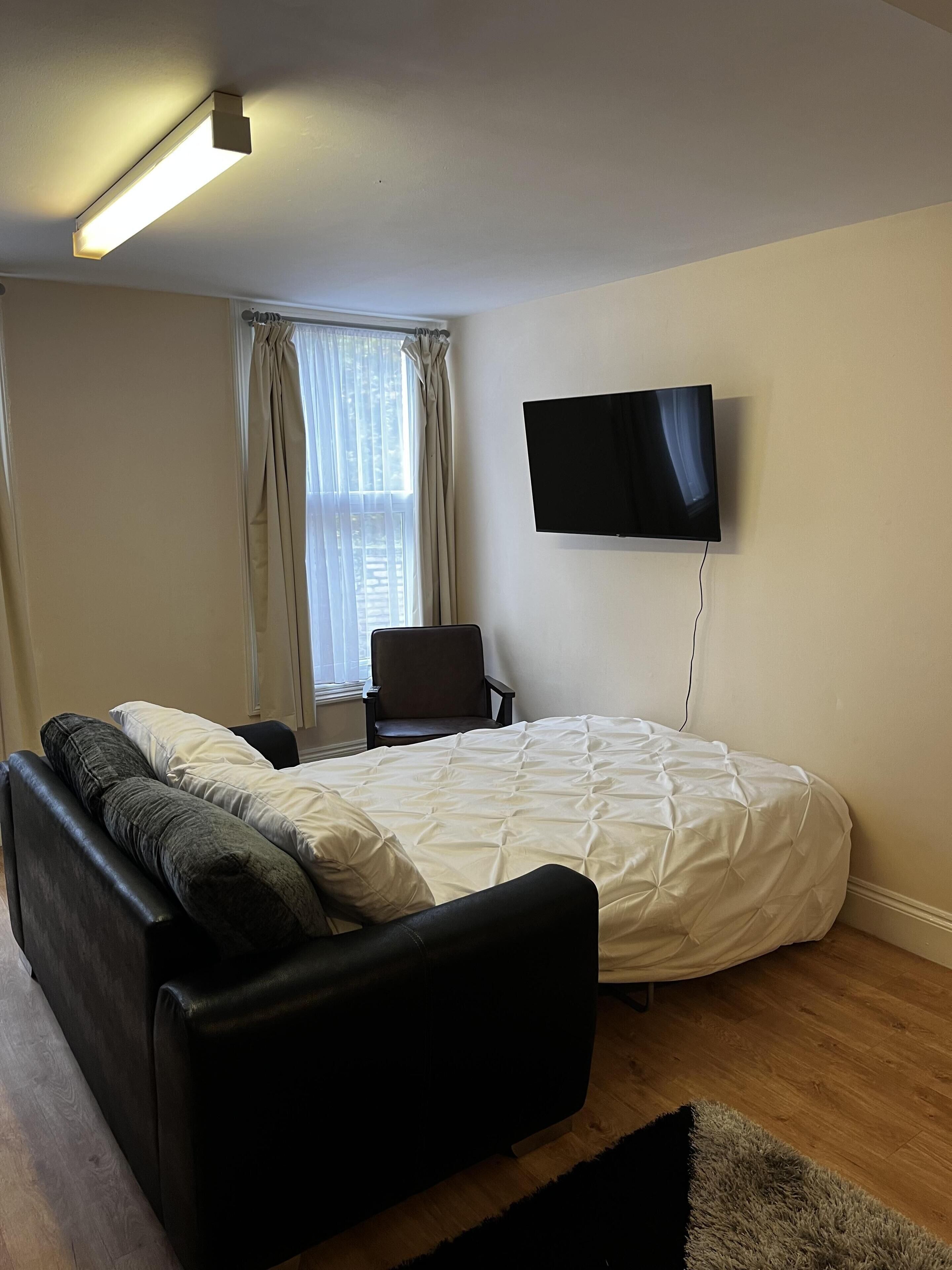 A bedroom with a bed, a sofa, a chair, a TV, and a window with curtains.