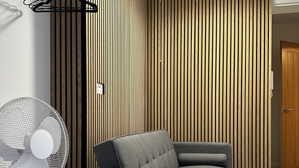 A modern room with a grey sofa, a standing fan, and a wooden slatted wall. - Flexible One Bedroom Apartment With Sofa Beds (Colchester)