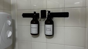 A bathroom with white tiles, a white hand dryer, and two bottles of shampoo and body wash hanging on a black rack. - Flexible One Bedroom Apartment With Sofa Beds (Colchester)