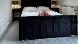A modern bedroom with a large bed, black headboard, and a window with a view of trees. There are two bedside tables with lamps, and a radiator on the wall. - Flexible One Bedroom Apartment With Sofa Beds (Colchester)