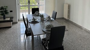 A modern dining area with a rectangular table set for four, surrounded by chairs. There is a potted plant on the left, a radiator on the right, and a large window with a view of greenery outside. - Cozy apartment within walking distance of the sea, with pets allowed  (Scalo Ferroviario)
