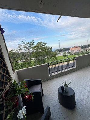 Un balcon donnant sur une rue et des immeubles, une chaise noire, une petite table avec un pot de plante et des fleurs en pot.
