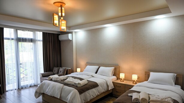 A hotel room with two beds, a sofa, a large window with curtains, and a ceiling light fixture. - K124 (Kutaisi)