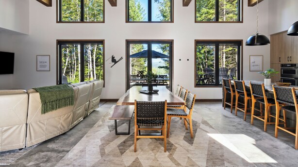 Un vaste espace de salle à manger avec une longue table et des chaises en bois, de grandes fenêtres donnant sur la verdure, et une cuisine moderne en arrière-plan.