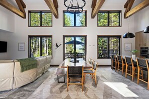 Uma sala de jantar ampla com uma longa mesa de madeira e cadeiras, grandes janelas com vista para árvores, e uma cozinha moderna ao fundo.