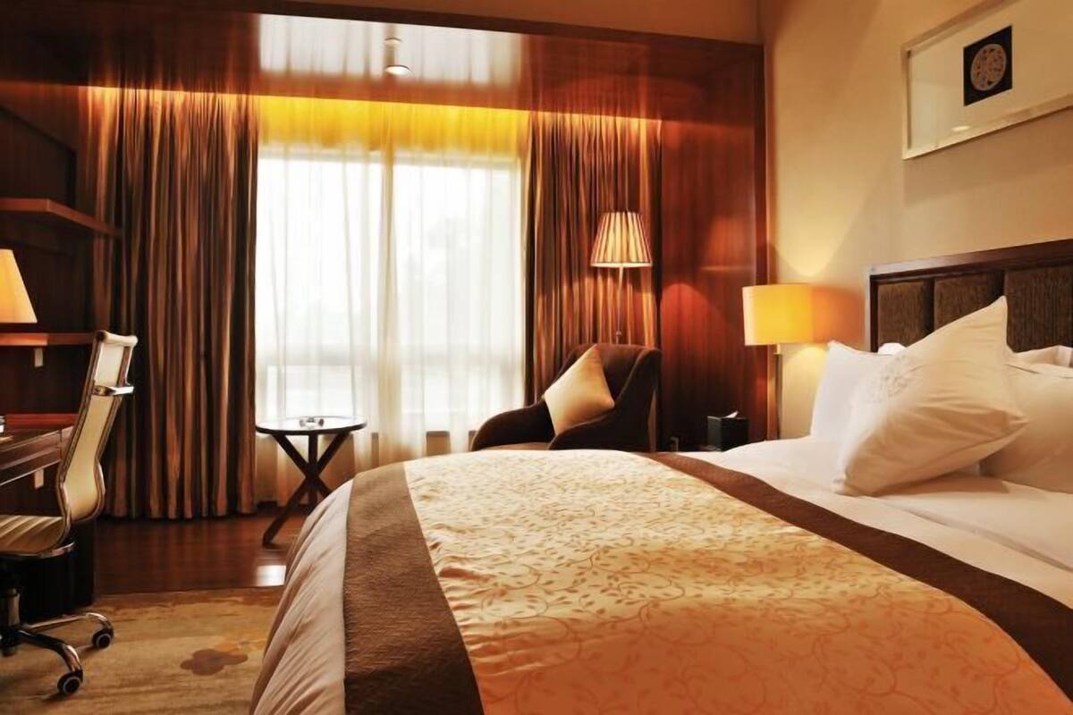 A hotel room with a large bed, a desk, a chair, a lamp, and a view through the window.
