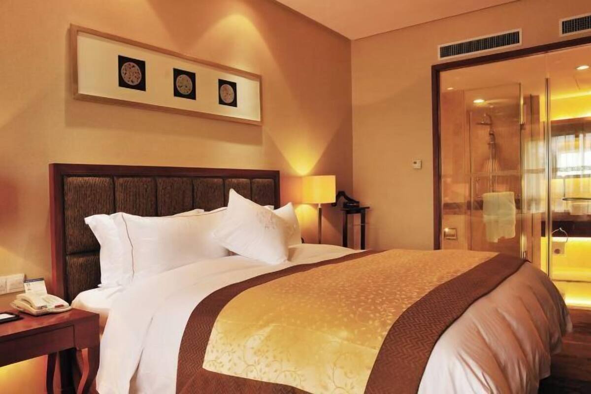 A hotel room with a large bed, a bedside table with a lamp, a glass shower enclosure, and framed pictures on the wall.