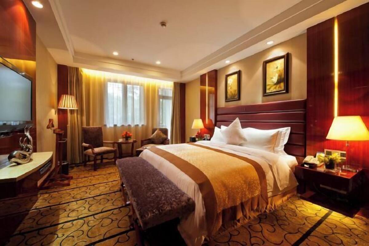 A hotel room with a large bed, a desk, a chair, a bench, a TV, and a window with curtains.