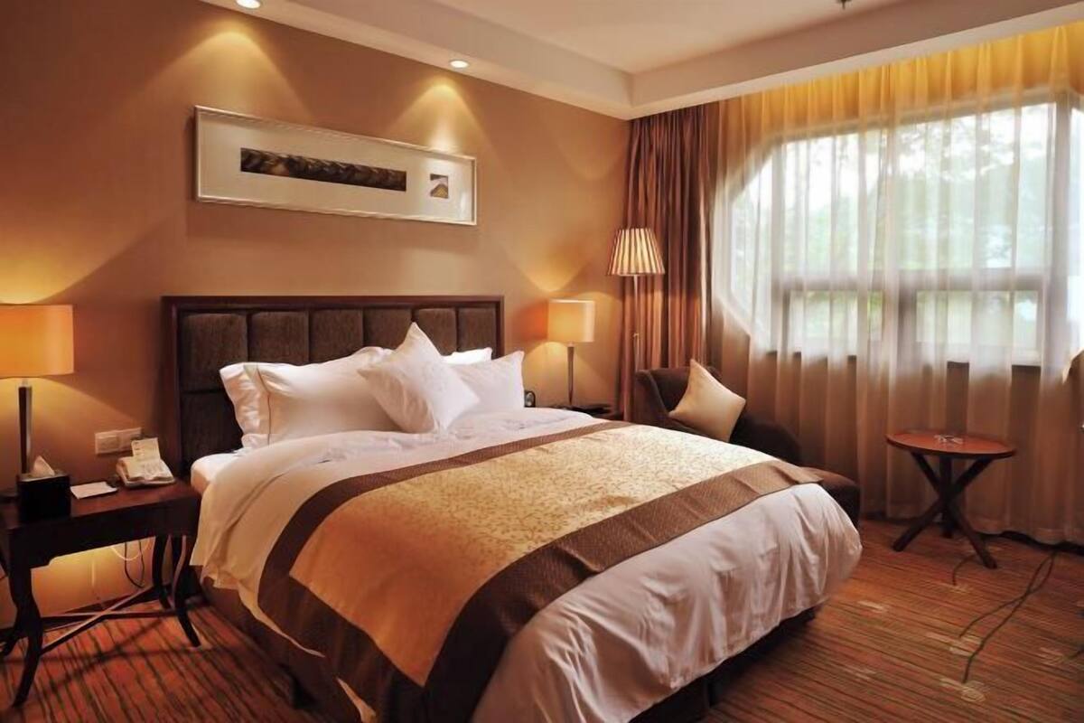A hotel room with a large bed, bedside lamps, a small table, and a window with curtains.