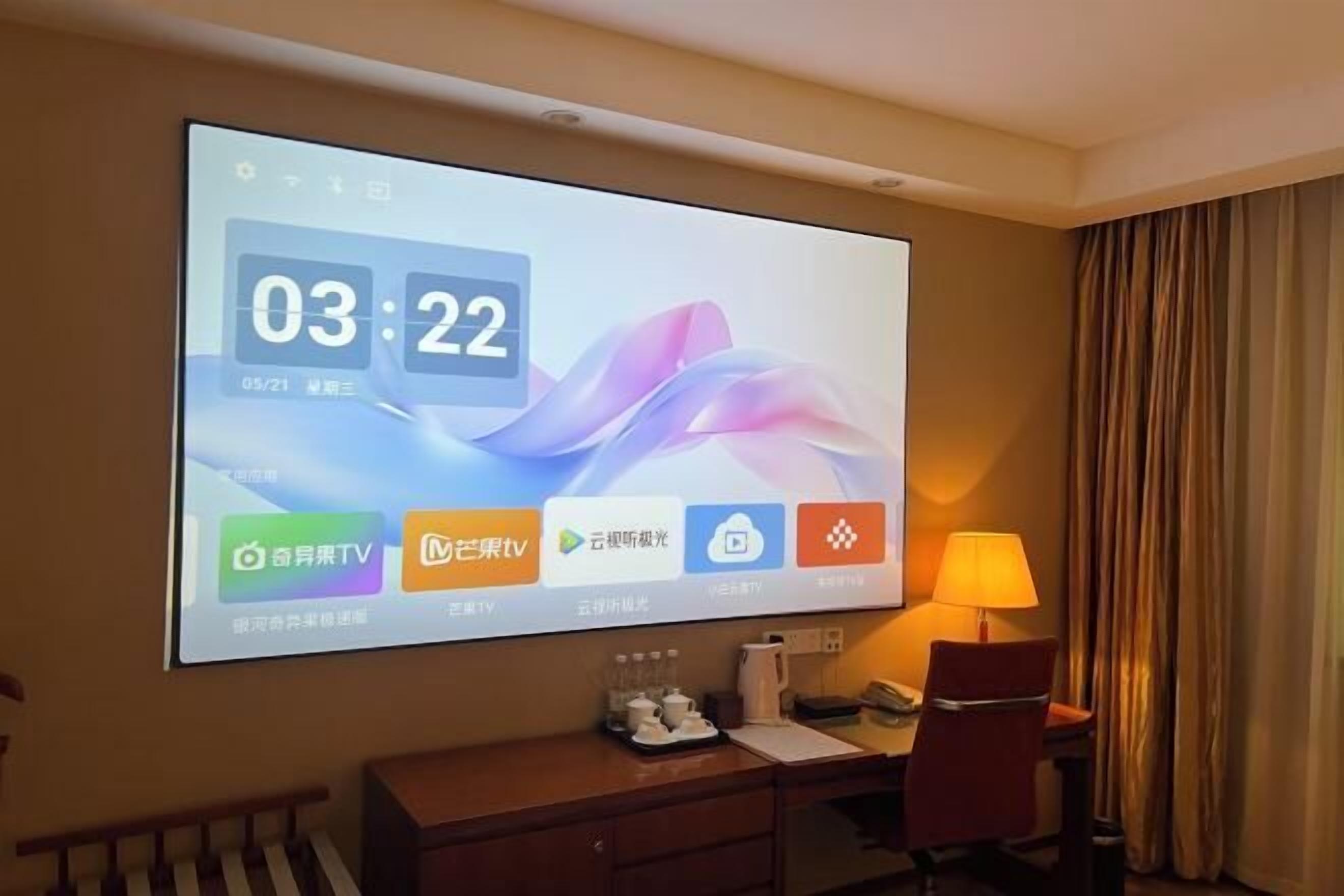 A hotel room with a large digital display on the wall, a desk with a lamp, and a chair.