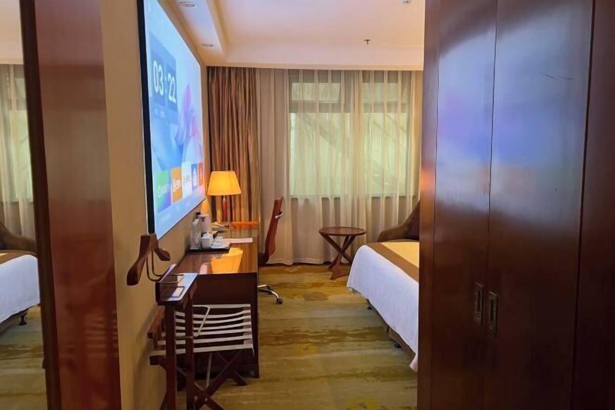 A hotel room with a bed, a desk with a lamp, a chair, a small table, and a projector screen displaying a presentation.