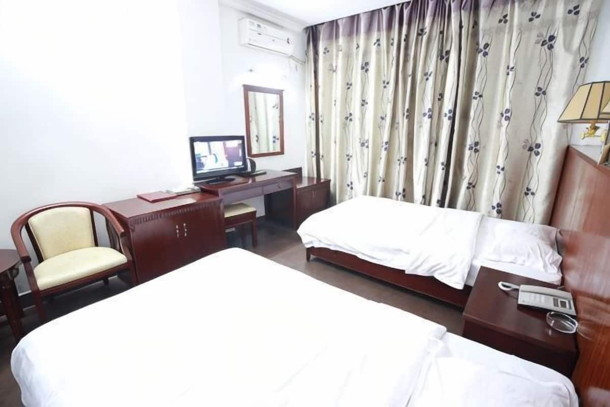 A hotel room with a bed, a desk with a computer, a chair, and a bedside table with a lamp.