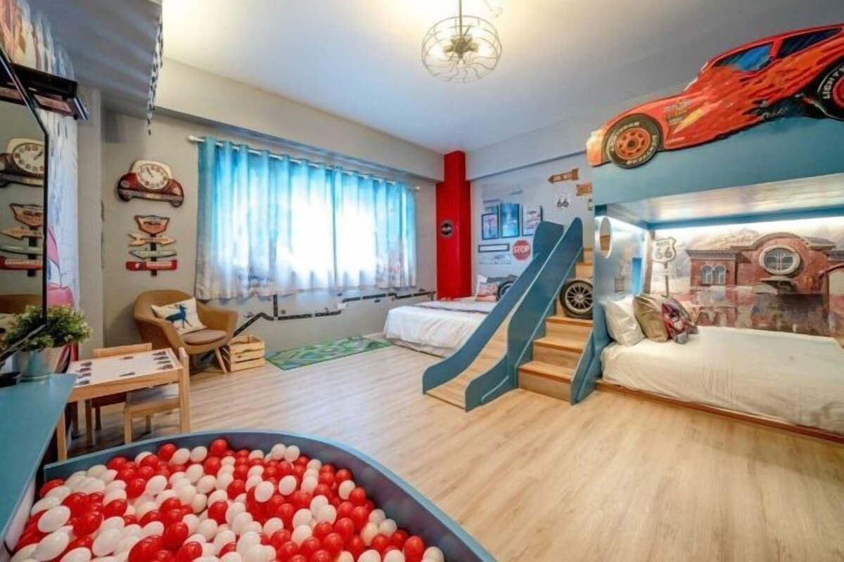 Une chambre d’enfant avec un lit, un espace de jeux comprenant une piscine à balles et un toboggan, ainsi qu’un mur décoré sur le thème des voitures et des courses automobiles.