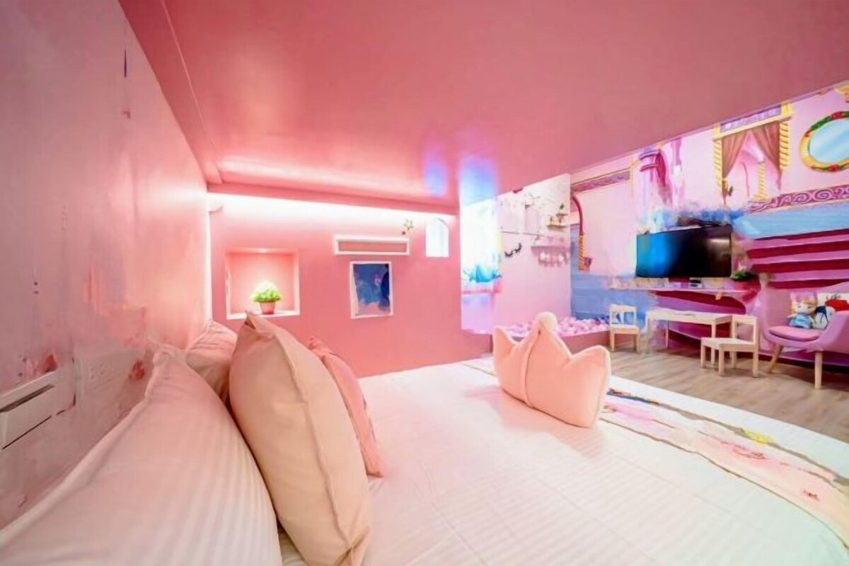 Une chambre à coucher décorée dans des tons roses, avec un lit, des oreillers, une télévision et un petit bureau.