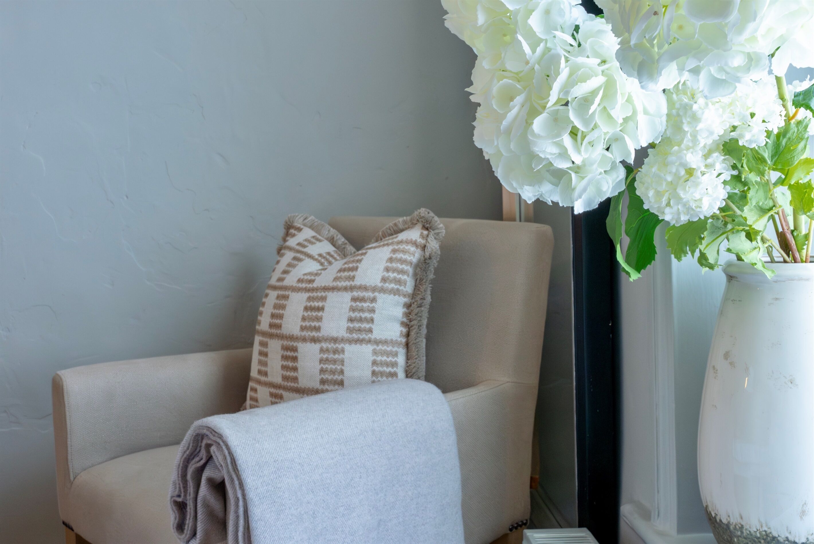 Une chaise avec un coussin à motifs et une couverture, un vase blanc avec des fleurs et un mur uni.