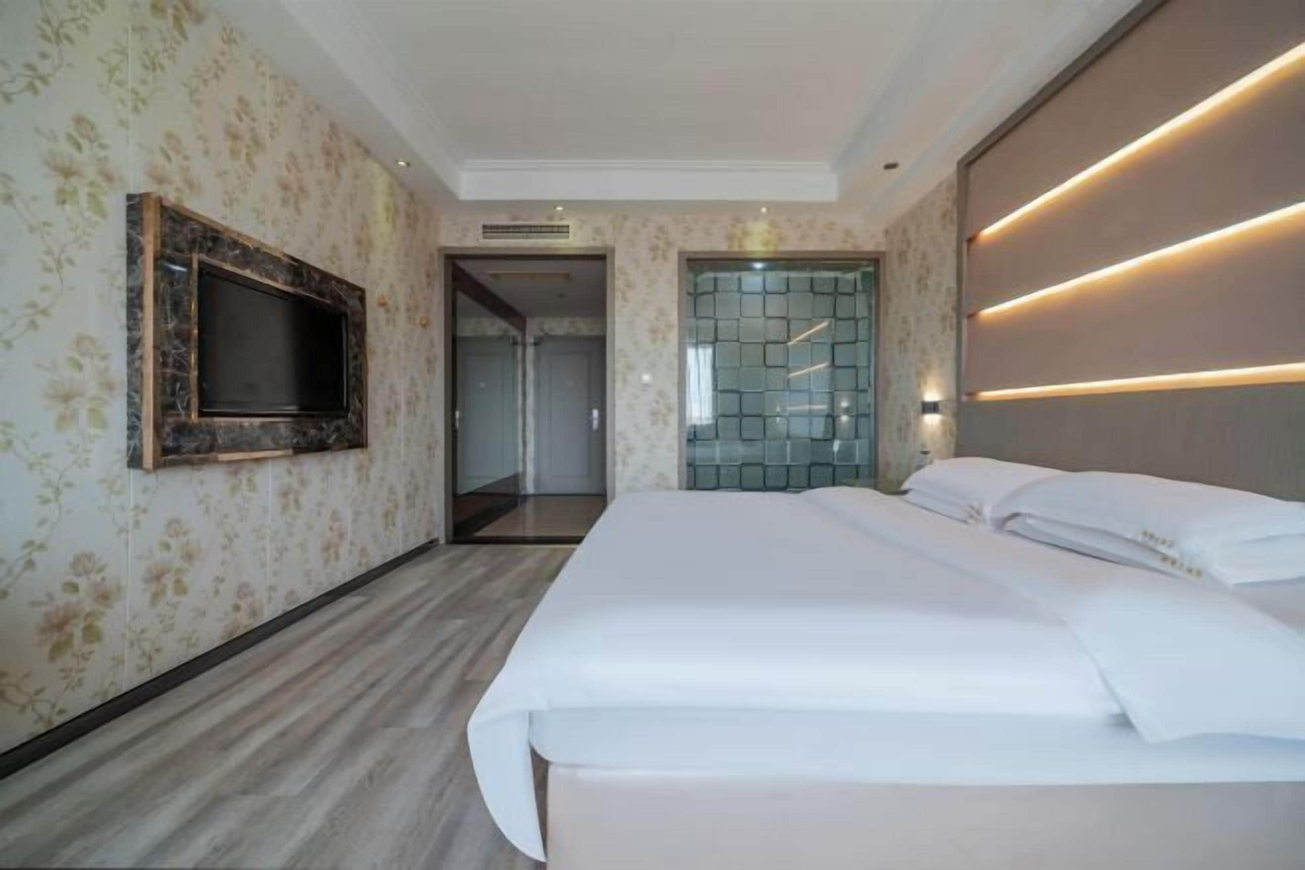 Une chambre d’hôtel moderne dotée d’un grand lit, d’un téléviseur à écran plat fixé au mur et d’une porte en verre donnant sur la salle de bain.