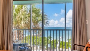 Ein Zimmer mit einer großen SchiebeglasTür, die auf einen Balkon mit Blick auf das Meer und Palmen führt. Auf dem Balkon stehen Korbsessel und ein kleiner Tisch.