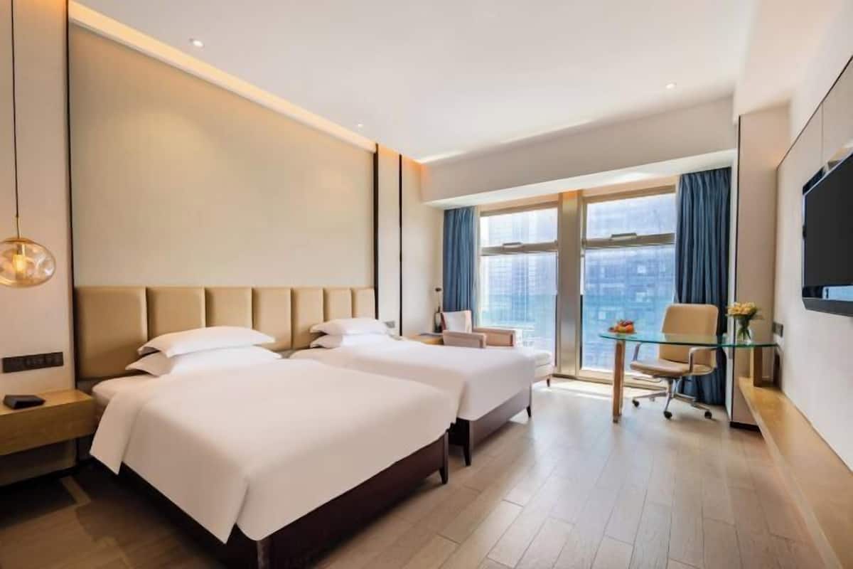 A hotel room with two beds, a desk, a chair, a TV, and a large window with a city view.